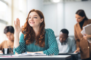 Eine Frau sitzt während einer Fortbildung an einem Tisch und hebt die Hand, um sich zu melden.