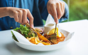 Nahaufnahme der Hände einer Person, die Besteck halten und die Speise auf dem Teller vor ihr Schneiden. Es sind Pommes Frites und Salat zu sehen.